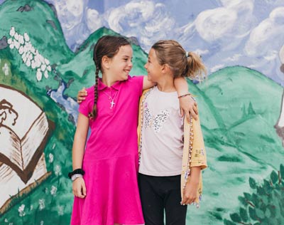 Two young girls with arms around each other smiling in front of a mural